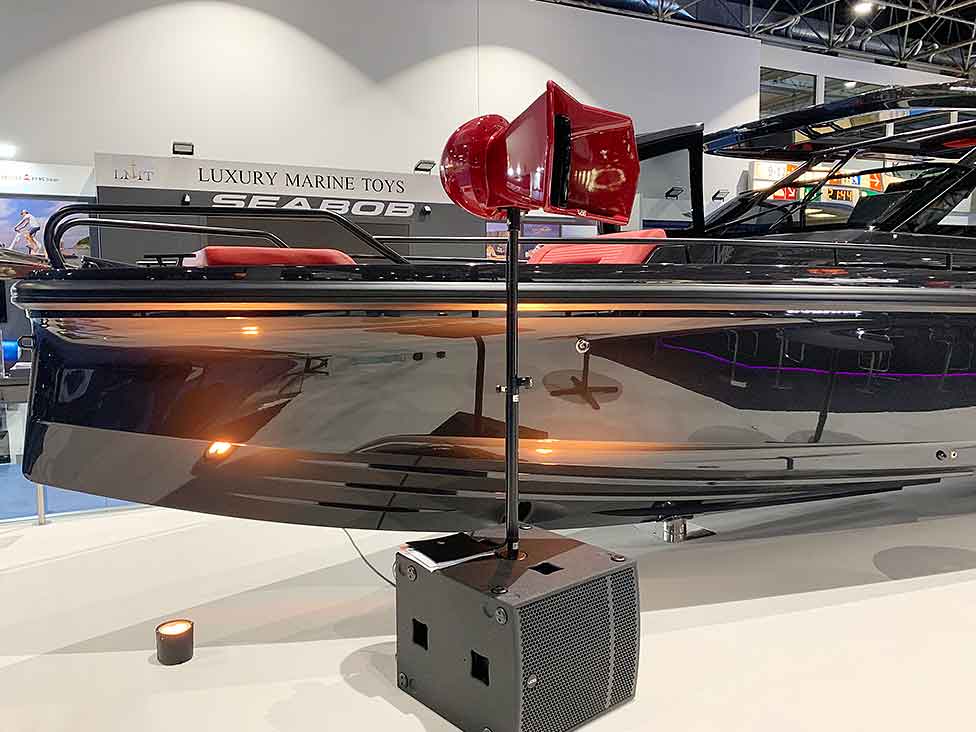 Void Acoustics Arcline 118 Subwoofer und rotes Hornlautsprechersystem auf einer Messe, platziert vor einem luxuriösen schwarzen Boot Void Acoustics Arcline 118 Subwoofer und rotes Hornlautsprechersystem auf einer Messe, platziert vor einem luxuriösen schwarzen Boot